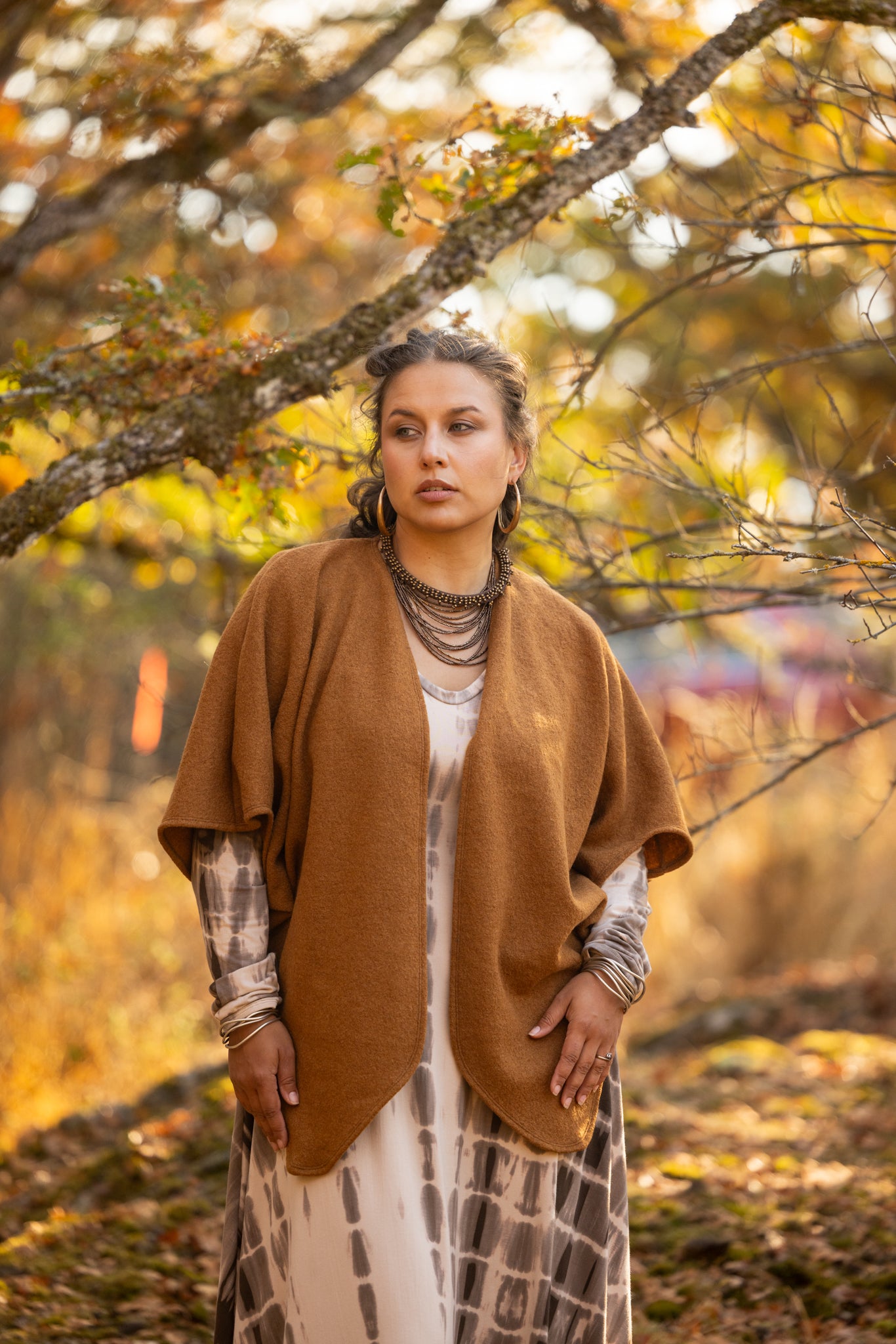 A woman stands outdoors in a wooded, autumn setting wearing a brown open-front poncho over a white and gray patterned dress. The background features soft-focus trees with yellow and orange foliage.